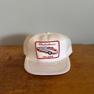 May include: A white trucker cap featuring a rectangular patch on the front. The patch displays a red and white sports car illustration with the text Charterhouse above the car and the word COLLISION printed in red below the vehicle graphic.