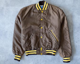 May include: A brown satin bomber jacket with yellow and black striped trim on the collar, cuffs, and hem. The jacket has yellow snap closures and the name "Ernest Bowman" embroidered in gold on the front.