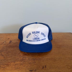 May include: A blue and white trucker hat featuring the text "INTERIOR BUILDING SUPPLIES LONDON" in blue. The hat has a blue peak and a white front panel with the company's logo. The hat is on a wooden surface.