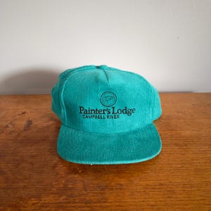 May include: A teal corduroy baseball cap with a curved peak. The cap displays the text "Painter's Lodge Campbell River" in black, and a circular logo. This classic hat is suitable for casual wear.