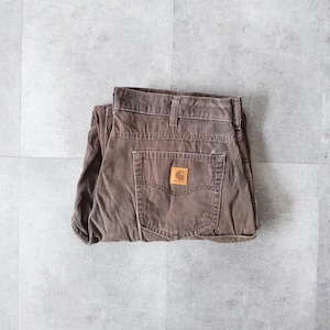 Puede incluir: Pantalones de trabajo Carhartt marrones doblados sobre una superficie gris. Los pantalones tienen un dise&ntilde;o cl&aacute;sico de cinco bolsillos, un parche de cuero marr&oacute;n con el logotipo de Carhartt y un aspecto desgastado. Fabricados con tejido duradero y resistente.