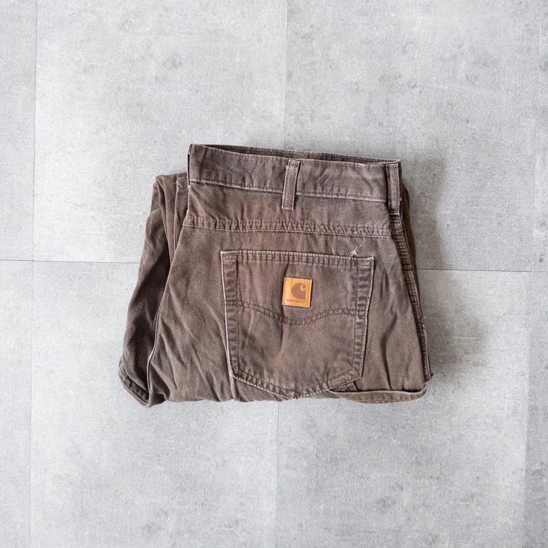 Puede incluir: Pantalones de trabajo Carhartt marrones doblados sobre una superficie gris. Los pantalones tienen un dise&ntilde;o cl&aacute;sico de cinco bolsillos, un parche de cuero marr&oacute;n con el logotipo de Carhartt y un aspecto desgastado. Fabricados con tejido duradero y resistente.