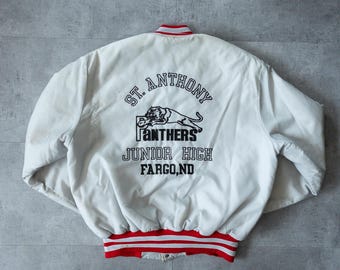 May include: White varsity jacket with red and white striped trim. The back features black text and a panther graphic that reads "St. Anthony Junior High, Fargo, ND". The jacket has a ribbed collar and cuffs.