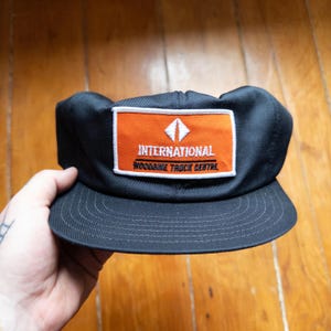 May include: Black baseball cap with a flat peak. The cap has an orange rectangular patch with the word "INTERNATIONAL" above "WOODBINE TRUCK CENTRE" in white lettering. A white diamond shape is above the word "INTERNATIONAL".