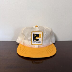 May include: A yellow and white mesh trucker hat featuring a rectangular patch on the front. The patch displays a black and yellow graphic logo with the text KAR PRODUCTS printed in black capital letters below the design. The hat has a solid yellow brim.