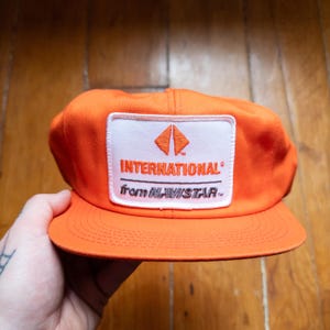 May include: A bright orange baseball cap with a white patch displaying the word "INTERNATIONAL" and the phrase "from NAVISTAR". The cap has a curved peak and a classic six-panel design. A vibrant accessory.