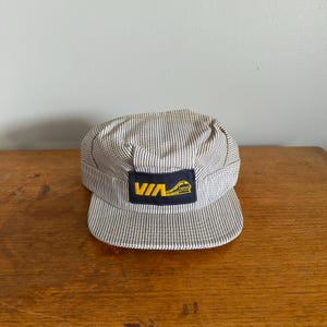 May include: A striped engineer's cap with a small brim. The cap is white and grey with vertical stripes. A navy blue patch on the front displays the word "VIA" in yellow, along with a yellow train graphic. The cap sits on a wooden surface.