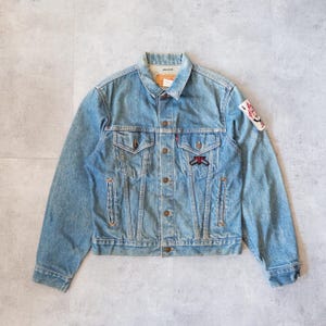 Puede incluir: Chaqueta vaquera azul claro con botones, dos bolsillos en el pecho y bolsillos laterales. Cuenta con parches en las mangas. La chaqueta tiene un cuello clásico y un aspecto vintage.