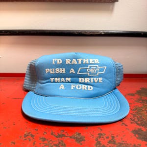 May include: A light blue trucker hat featuring a mesh back and a foam front panel. The front displays white embroidered text that reads I'D RATHER PUSH A CHEVY THAN DRIVE A FORD, including the Chevrolet logo between the words PUSH A and THAN.