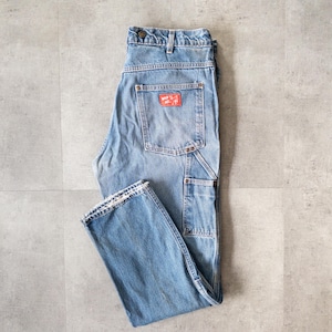 Può includere: Pantaloni da lavoro in denim blu chiaro con più tasche e una toppa rossa sulla tasca posteriore. I pantaloni hanno un aspetto classico e usurato con chiusura con bottoni e cerniera. L'orlo inferiore è sfilacciato.