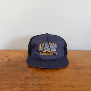 Op de afbeelding: Een marineblauwe truckerpet met een zwarte mesh achterkant en een platte klep. Het voorpaneel toont geborduurde tekst in blauw met een gele omlijning die UAW leest, met de tekst LOCAL 222 in het geel binnen een rechthoekige rand eronder.
