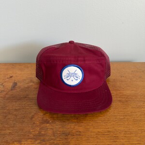 Può includere: Un berretto da baseball bordeaux con retro in rete e visiera curva. La parte anteriore del berretto presenta una toppa circolare con mazze da golf incrociate e la scritta "Baldoon Golf & Co" in blu e bianco.