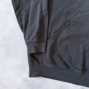 Puede incluir: Primer plano de una sudadera con capucha negra. La sudadera tiene un bolsillo delantero y pu&ntilde;os y dobladillo acanalados. La tela parece ser un material suave y c&oacute;modo.