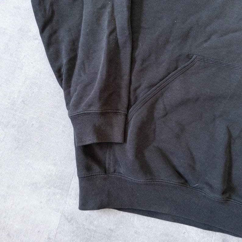 Puede incluir: Primer plano de una sudadera con capucha negra. La sudadera tiene un bolsillo delantero y pu&ntilde;os y dobladillo acanalados. La tela parece ser un material suave y c&oacute;modo.