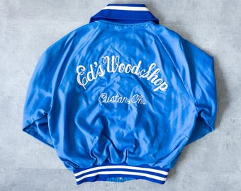 May include: A blue satin jacket with white trim and the text "Ed's Woodshop" embroidered on the back. The jacket has a collar and a striped hem. The text "Custar, Oh." is also embroidered on the back.