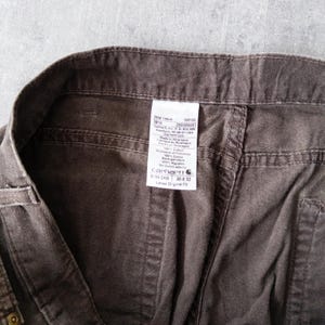 Puede incluir: Primer plano de unos pantalones de trabajo marrones Carhartt. La etiqueta indica una talla de 38 x 32 pulgadas (96,5 x 81,3 cm). Los pantalones est&aacute;n hechos de una tela duradera y presentan un dise&ntilde;o cl&aacute;sico de ajuste holgado. La etiqueta incluye la marca y otros detalles.