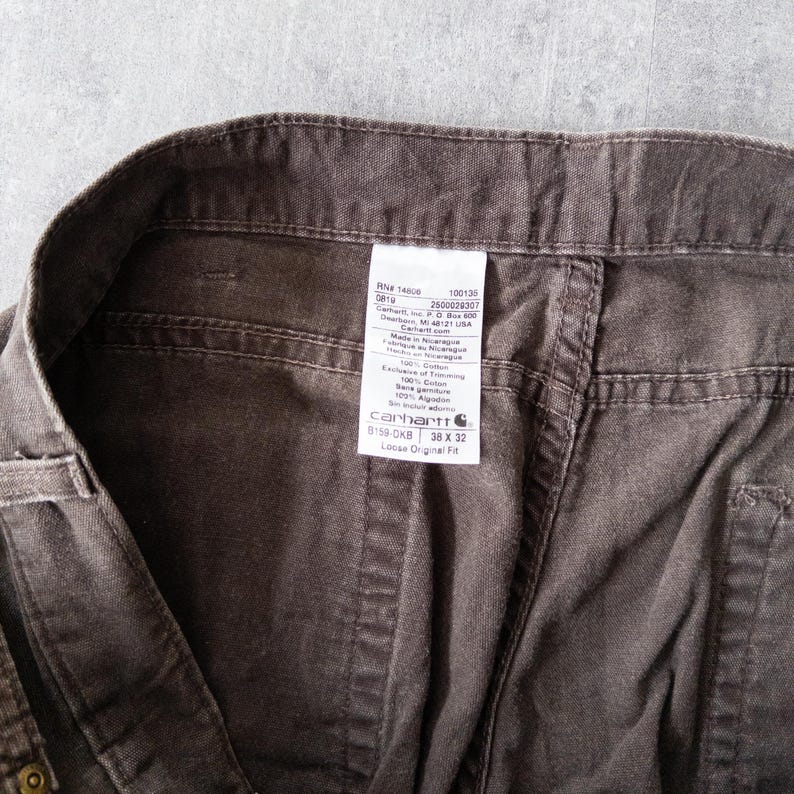 Puede incluir: Primer plano de unos pantalones de trabajo marrones Carhartt. La etiqueta indica una talla de 38 x 32 pulgadas (96,5 x 81,3 cm). Los pantalones est&aacute;n hechos de una tela duradera y presentan un dise&ntilde;o cl&aacute;sico de ajuste holgado. La etiqueta incluye la marca y otros detalles.