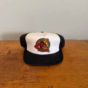 May include: A black and white trucker hat featuring a graphic on the front panel. The graphic depicts a red devil figure playing a yellow fiddle, surrounded by a green circular border with the text Golden Fiddle Music printed in gold lettering.