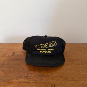 May include: A black trucker hat featuring a mesh back and a solid front panel. The front displays yellow screen-printed text that reads DA YOOPERS WORLD TOUR 1990. The cap has a structured crown, a curved brim, and an adjustable snapback closure.