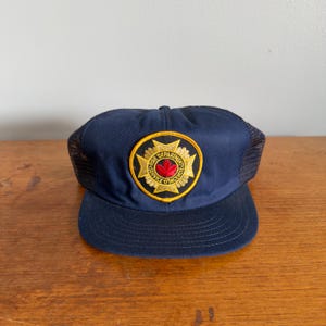 Puede incluir: Gorra de camionero azul marino con parte trasera de malla y visera curva. La parte delantera presenta un parche circular con ribete dorado, las palabras "FIRE DEPARTMENT SERVICE D'INCENDIE" y una hoja de arce roja.