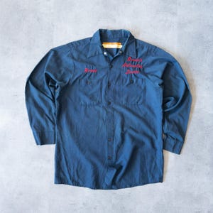 Może przedstawiać: Długoręczna koszula robocza w ciemnoniebieskim kolorze z dwiema kieszeniami na piersi i zapięciem na guziki. Koszula ma czerwony haft na prawej i lewej stronie piersi.