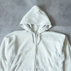 Pu&ograve; includere: Una felpa con cappuccio grigio chiaro con cerniera e cappuccio con coulisse. La felpa ha una cerniera a tutta lunghezza e una tasca frontale. Il tessuto sembra essere un materiale morbido e confortevole, adatto all'abbigliamento casual.