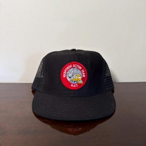 Peut inclure: Une casquette de camionneur noire avec un écusson circulaire sur le panneau avant. L'écusson a une bordure rouge et contient le texte RESPONSE ACTION TEAM et R.A.T. avec une illustration d'un rat gris conduisant un camion jaune sur fond bleu.