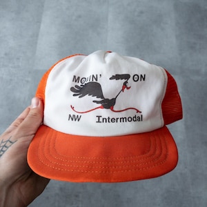 Può includere: Un cappello da camionista bianco e arancione con un design di roadrunner dei cartoni animati. Il cappello ha il testo "MOVIN' ON" e "NW Intermodal" sul davanti.