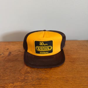 Peut inclure: Une casquette trucker marron et jaune avec un écusson rectangulaire noir sur le devant. L'écusson affiche le texte 30 yrs. RAYWAL kitchens brodé en jaune. La casquette possède un panneau avant jaune, des panneaux arrière en maille marron.
