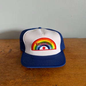 May include: A blue and white trucker hat with a rainbow and the words "Niagara Falls" embroidered on the front. The hat has a mesh back and a curved brim. The rainbow design features red, orange, yellow, green, and blue colors.