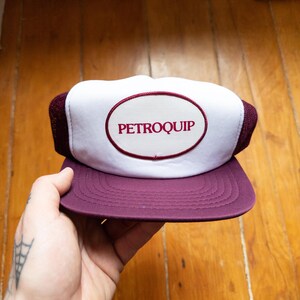 Op de afbeelding: Bordeauxrode en witte truckerpet met het woord "PETROQUIP" in rood geborduurd op een witte ovale patch. De pet heeft een bordeauxrode klep en een mesh achterkant. Waarschijnlijk een vintage promotieartikel.