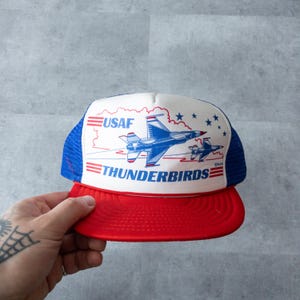 May include: A white, blue and red mesh trucker hat with the words "USAF THUNDERBIRDS" and a graphic of two fighter jets.