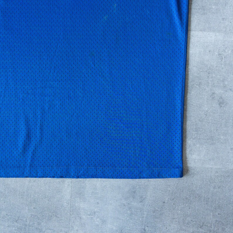 May include: Close-up of a vibrant blue mesh fabric, likely from a jersey or athletic wear. The material has a textured, perforated surface, and the color is a solid, bright blue. The fabric is neatly cut and sewn along the bottom edge.