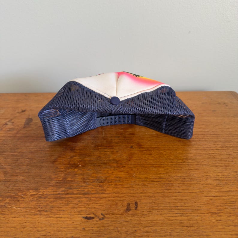 May include: A trucker hat with a white front panel featuring a colourful design, a navy blue mesh back, and a navy blue adjustable snap closure. The hat has a curved brim and a button on top.