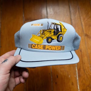Puede incluir: Gorra de camionero azul claro con un gráfico amarillo de retroexcavadora Case Power. La gorra presenta el texto "580K" y "CASE POWER" en negrita. La gorra tiene una visera negra y una parte trasera de malla.