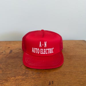 May include: A red trucker hat with the text "A · N AUTO ELECTRIC" in white. The hat has a mesh back and a curved brim. The hat is sitting on a wooden surface.
