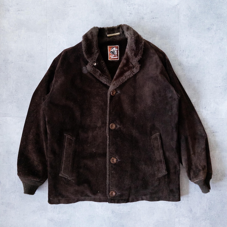 K&ouml;nnte beinhalten: Dunkelbraune Wildlederjacke mit Kunstfellkragen und -futter. Die Jacke hat eine Knopfleiste, zwei Vordertaschen und gerippte B&uuml;ndchen. Ein kleines Logo-Patch ist am Kragen sichtbar.
