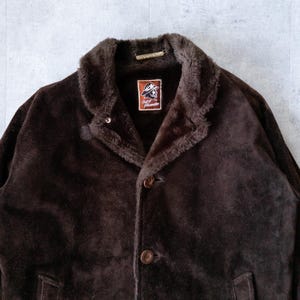 K&ouml;nnte beinhalten: Dunkelbraune Wildlederjacke mit Kunstfellkragen und -futter. Die Jacke hat einen Knopfverschluss und ein Etikett mit einem Logo. Die Jacke ist im Vintage-Stil und scheint in gutem Zustand zu sein.