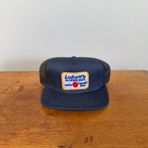 May include: A navy blue trucker hat with a black mesh back. The front of the hat features a rectangular patch with the text "Labatt's Pilsener Beer" and "call for Labatt's Blue". The patch has a yellow border and red and blue accents.
