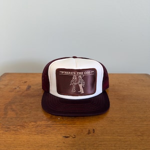 Könnte beinhalten: Burgunderfarbene und weiße Trucker-Mütze mit einem rechteckigen Aufnäher mit dem Text "WHERE'S THE COD ?" und einer Illustration von zwei Figuren. Die Mütze hat einen Mesh-Rücken und einen gebogenen Schirm.
