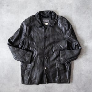 May include: Black leather jacket with a button-up front, two front pockets, and a collared neckline. The jacket has a classic design and a slightly worn appearance. The brand name "COSA NOVA" is visible on the label.