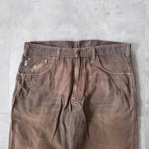 Puede incluir: Primer plano de pantalones de trabajo marrones con aspecto descolorido. Los pantalones tienen cierre de bot&oacute;n, trabillas para el cintur&oacute;n y m&uacute;ltiples bolsillos. La tela muestra signos de desgaste, d&aacute;ndole un aspecto vintage. Los pantalones est&aacute;n hechos de un material duradero.