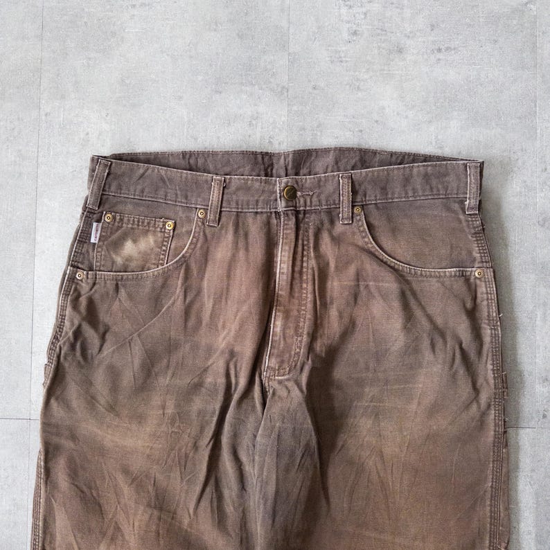 Puede incluir: Primer plano de pantalones de trabajo marrones con aspecto descolorido. Los pantalones tienen cierre de bot&oacute;n, trabillas para el cintur&oacute;n y m&uacute;ltiples bolsillos. La tela muestra signos de desgaste, d&aacute;ndole un aspecto vintage. Los pantalones est&aacute;n hechos de un material duradero.