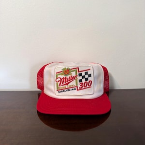 Puede incluir: Una gorra tipo camionero roja y blanca con un parche rectangular en la parte delantera. El parche muestra el logotipo de Miller High Life, un patrón de bandera a cuadros en blanco y negro, y el texto Miller High Life Syracuse N.Y. 300 en rojo.