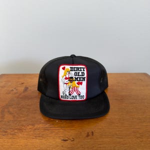 May include: Black trucker hat with a mesh back and a curved brim. The front patch features the text "DIRTY OLD MEN NEED LOVE TOO" with a cartoon illustration of two women and a man.