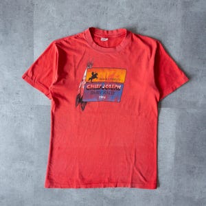 May include: Red short-sleeved t-shirt with a vintage graphic print of a Native American riding a horse with the text "BMW Presents Chief Joseph BMW Rally 1984".