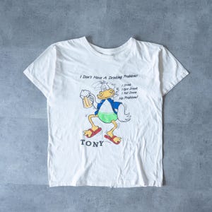 May include: White t-shirt featuring a cartoon duck holding a beer mug, wearing a blue jacket and green shorts, and red sandals. Text on the shirt reads "I Don't Have A Drinking Problem!" and "TONY".