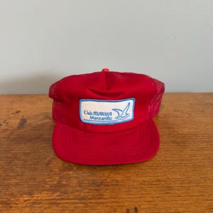May include: A red trucker cap featuring a mesh back and a solid front panel. A rectangular white patch is centred on the front, displaying the blue text Club MAEVA Manzanillo and a stylised blue bird graphic with a light blue border.