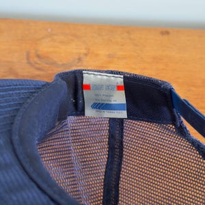 May include: A navy blue trucker cap with a mesh back and a curved peak. The cap has a tag with the text "SAN SUN" and other details. The cap is made of 100% polyester and is one size fits all.