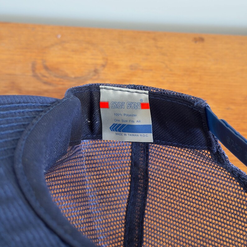 May include: A navy blue trucker cap with a mesh back and a curved peak. The cap has a tag with the text "SAN SUN" and other details. The cap is made of 100% polyester and is one size fits all.